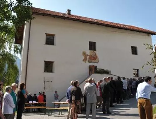 Eine Gruppe von Menschen steht in einer Reihe vor einem großen, weißen Gebäude. Im Hintergrund ist eine Wandmalerei zu sehen und es ist sonnig.