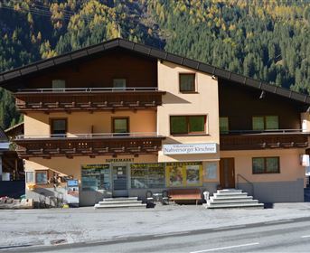 A cozy two-story building in the mountains with a supermarket on the ground floor. Surrounded by green forests and sunny weather.