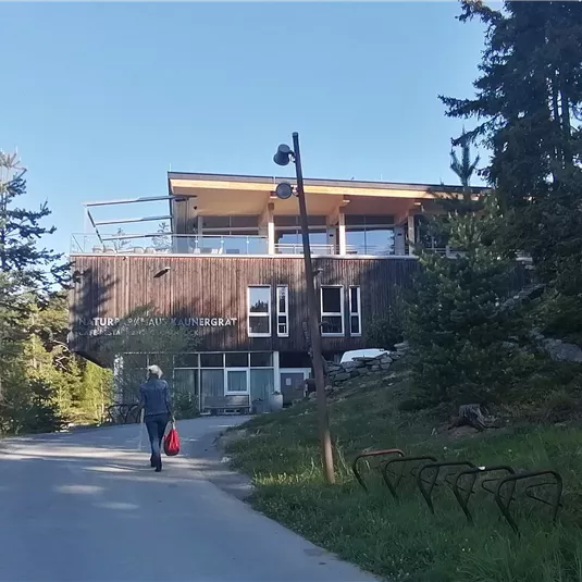 Ein moderner Holzbau steht am Ende eines schmalen Weges. Eine Person geht in Richtung des Gebäudes, umgeben von Bäumen und Grün.