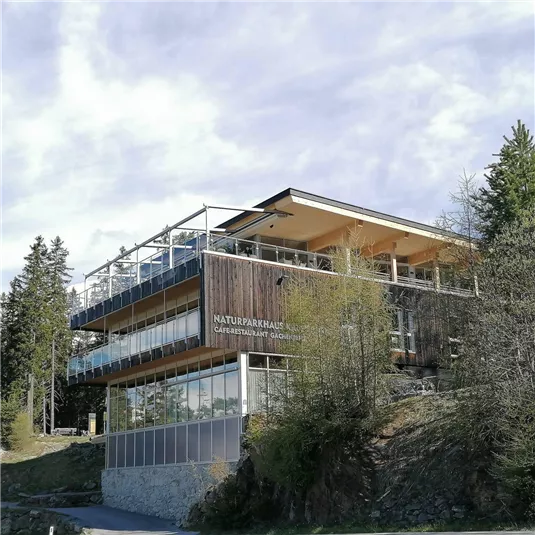 Ein modernes Gebäude mit Holz- und Glaselementen. Es steht in einer bewaldeten Umgebung und hat eine Terrasse mit Blick auf die Natur.