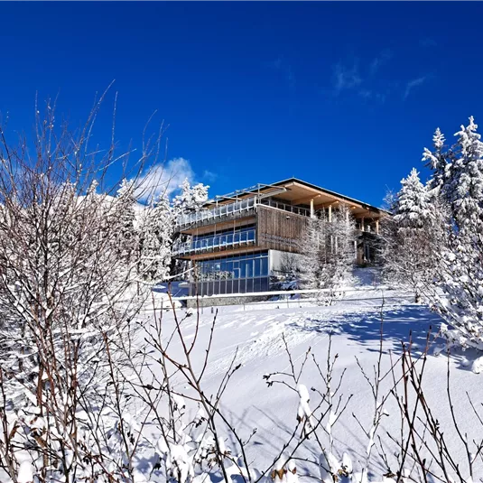 Ein modernes Gebäude steht in einer schneebedeckten Landschaft. Der klare blaue Himmel und die verschneiten Bäume schaffen eine ruhige Winteratmosphäre.