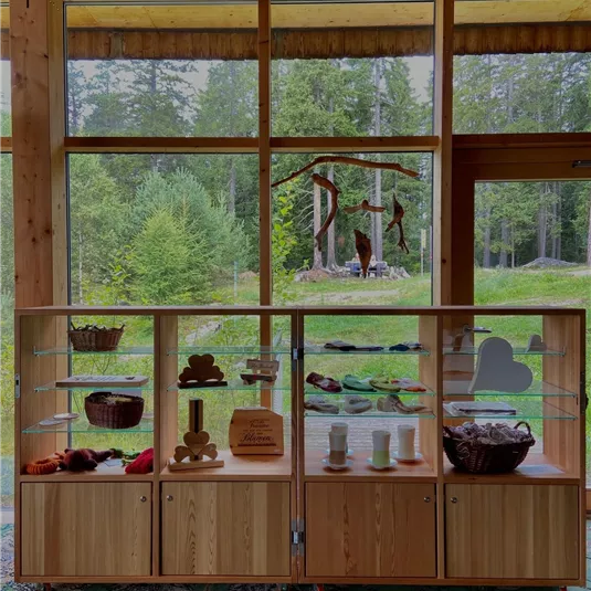 Ein Holzregal mit verschiedenen Körben und Dekorationsgegenständen steht vor großen Fenstern mit Blick auf eine grüne Landschaft. Die natürliche Beleuchtung schafft eine einladende Atmosphäre.