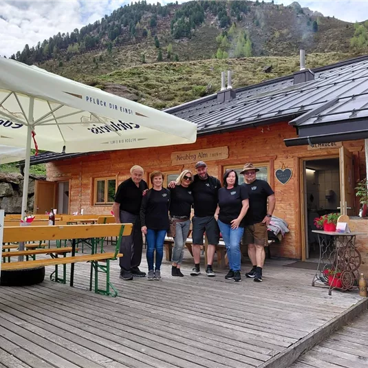 Eine Gruppe von sechs Personen steht vor einem Holzhaus in den Bergen. Es gibt Tische im Freien und eine Sonnenschirme.