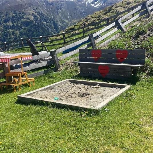 Eine gemütliche Sitzecke mit einer Bank und einem Sandkasten auf einer schönen Wiese in den Bergen. Im Hintergrund sind grüne Wiesen und Gehölze zu sehen.