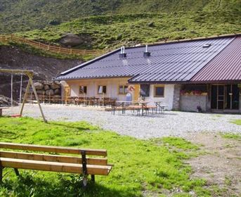 Ein gemütliches Berggasthaus mit einer Terrasse und Tischen im Freien. Im Vordergrund befindet sich eine Bank und eine Schaukel auf grünem Rasen.