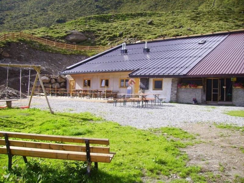 Ein gemütliches Berggasthaus mit einer Terrasse und Tischen im Freien. Im Vordergrund befindet sich eine Bank und eine Schaukel auf grünem Rasen.