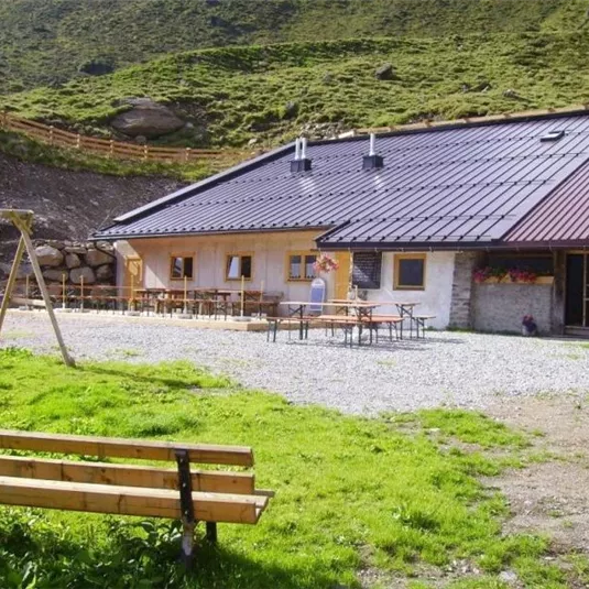 Ein gemütliches Berggasthaus mit einer Terrasse und Tischen im Freien. Im Vordergrund befindet sich eine Bank und eine Schaukel auf grünem Rasen.