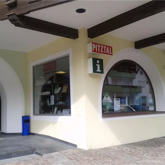 An information center in Pitztal with a large window and an inviting entrance area. The facade is light yellow, and a blue waste bin stands in front of the building.