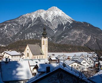 Eine winterliche Landschaft mit einem hohen, schneebedeckten Berg im Hintergrund. Im Vordergrund sind traditionelle Häuser und eine Kirche zu sehen.