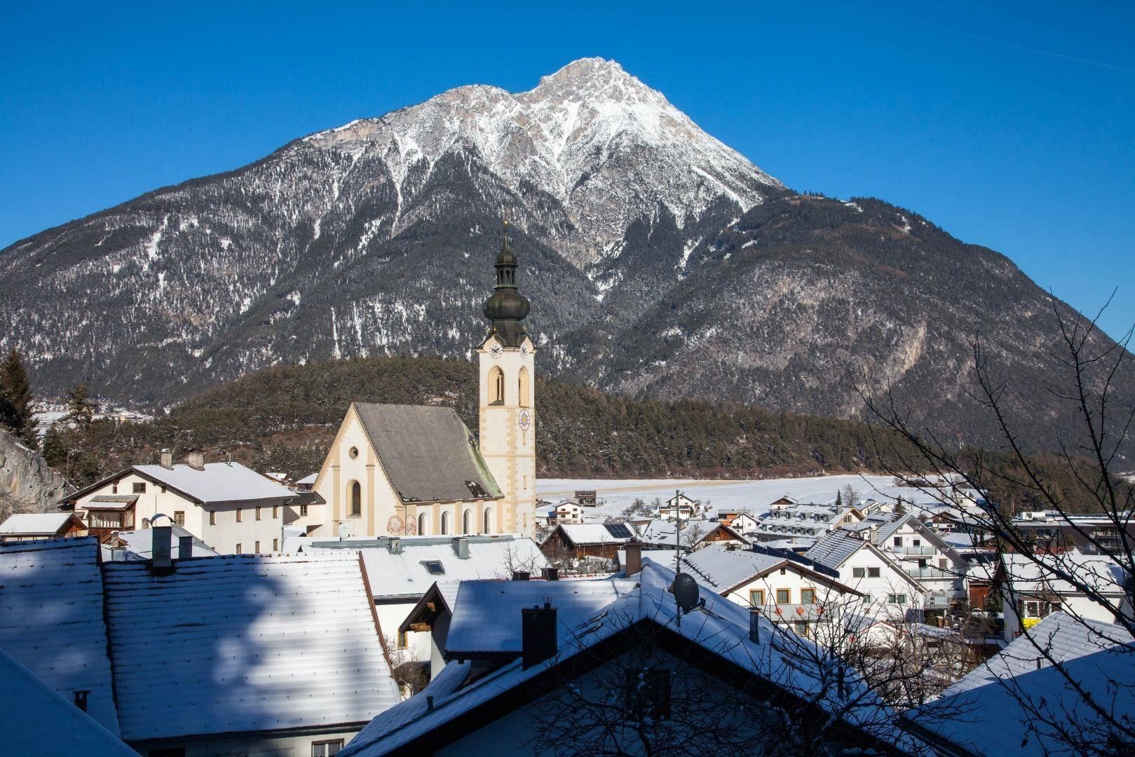 Eine winterliche Landschaft mit einem hohen, schneebedeckten Berg im Hintergrund. Im Vordergrund sind traditionelle Häuser und eine Kirche zu sehen.
