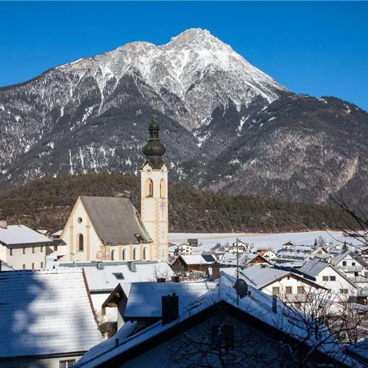 Eine winterliche Landschaft mit einem hohen, schneebedeckten Berg im Hintergrund. Im Vordergrund sind traditionelle Häuser und eine Kirche zu sehen.