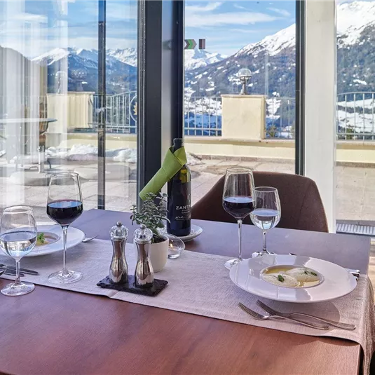 An elegant table with two place settings, wine, and a delicious dish. In the background, snow-capped mountains and a clear sky can be seen.