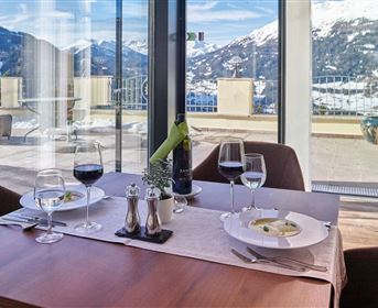 An elegant table with two place settings, wine, and a delicious dish. In the background, snow-capped mountains and a clear sky can be seen.