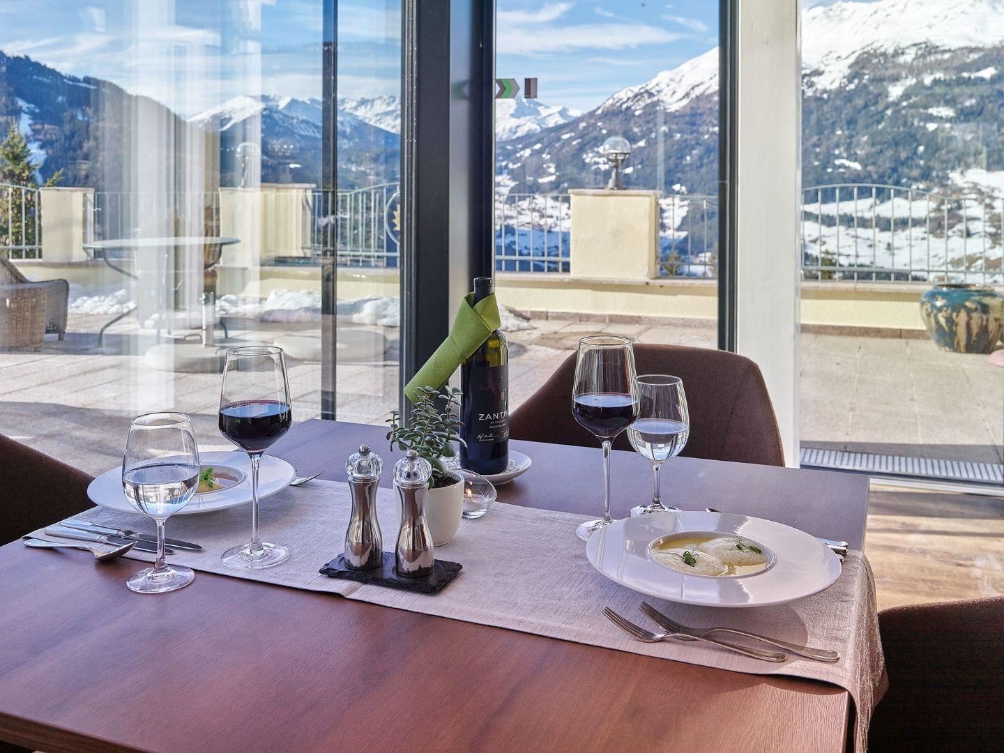 An elegant table with two place settings, wine, and a delicious dish. In the background, snow-capped mountains and a clear sky can be seen.