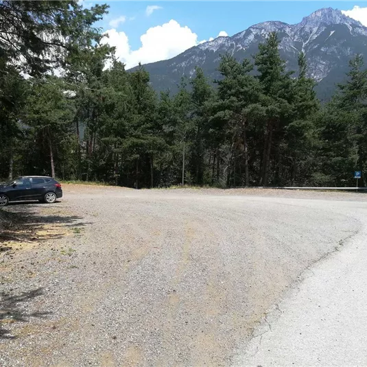 Ein Parkplatz umgeben von Bäumen und Bergen. Die Straße führt in eine bergige Landschaft mit klarem Himmel.