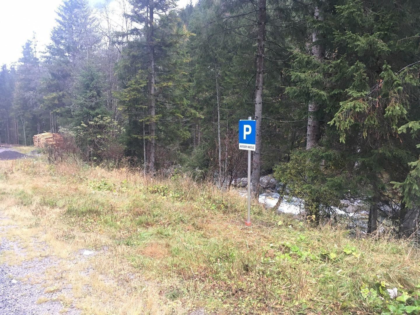 Ein Parkplatzschild steht neben einem schmalen Weg in einem Waldgebiet. Im Hintergrund fließt ein kleiner Bach zwischen den Bäumen.