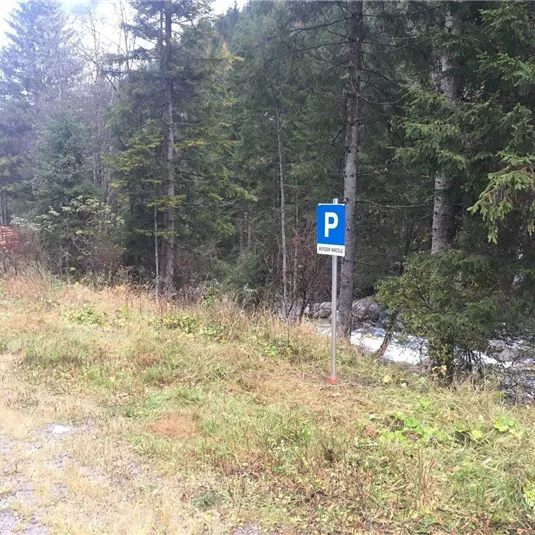 Ein Parkplatzschild steht neben einem schmalen Weg in einem Waldgebiet. Im Hintergrund fließt ein kleiner Bach zwischen den Bäumen.