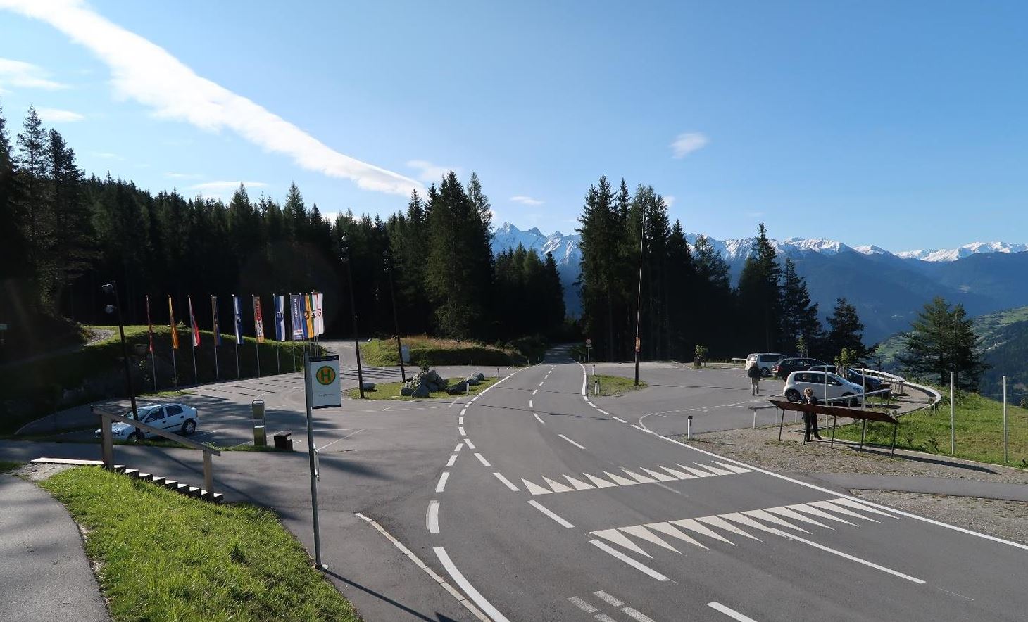 Ein malerischer Aussichtspunkt in den Bergen mit einer klaren Straße und grünen Wiesen. Im Hintergrund sind schneebedeckte Gipfel und Wälder zu sehen.