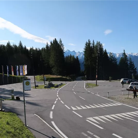 Ein malerischer Aussichtspunkt in den Bergen mit einer klaren Straße und grünen Wiesen. Im Hintergrund sind schneebedeckte Gipfel und Wälder zu sehen.