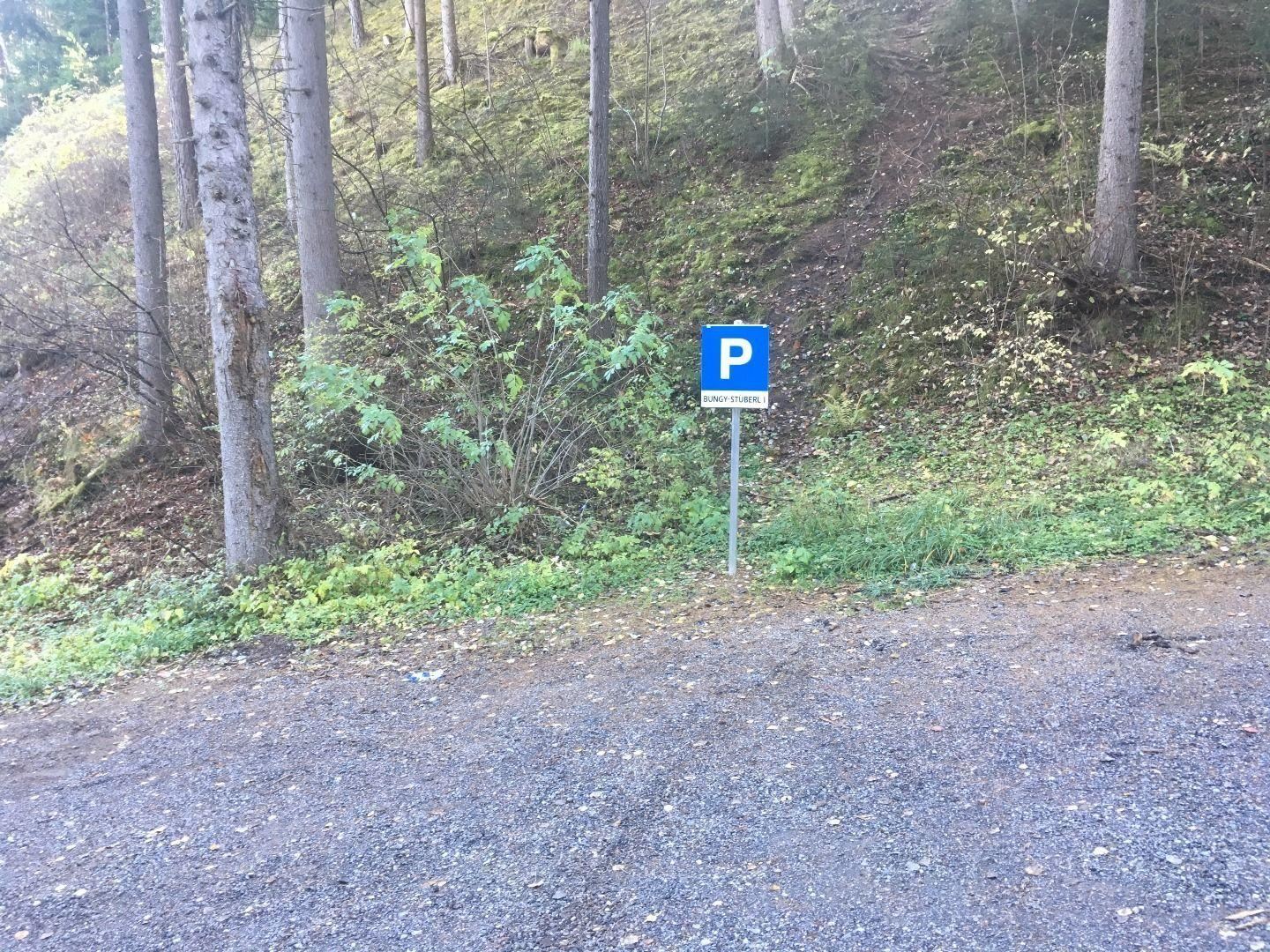 Ein Parkplatzschild steht an einem Waldweg. Die Umgebung ist grün und bewaldet.