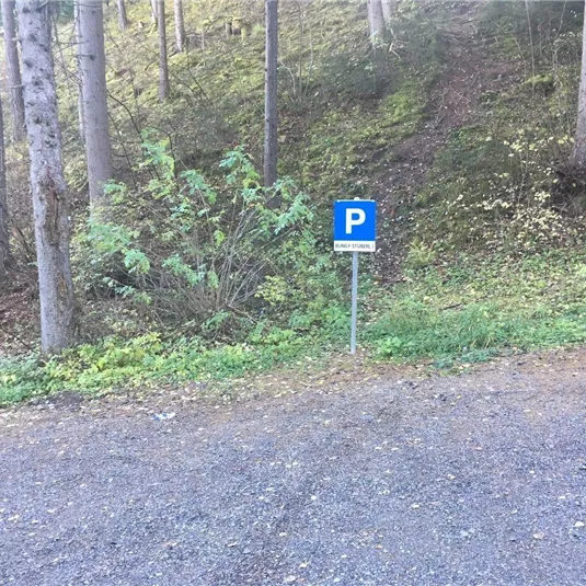 Ein Parkplatzschild steht an einem Waldweg. Die Umgebung ist grün und bewaldet.
