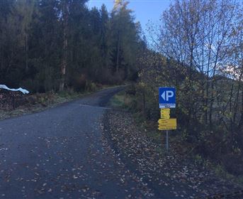 Ein Parkplatzschild zeigt die Richtung für Fahrzeuge. Der Weg führt durch eine bewaldete Umgebung mit herbstlichen Blättern.