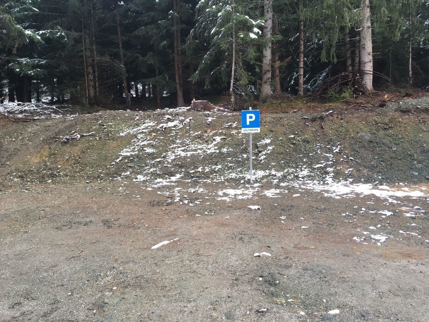Ein Parkplatzsschild steht an einer schneebedeckten Waldlichtung. Im Hintergrund sind grüne Bäume zu sehen.