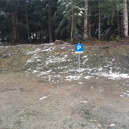 Ein Parkplatzsschild steht an einer schneebedeckten Waldlichtung. Im Hintergrund sind grüne Bäume zu sehen.