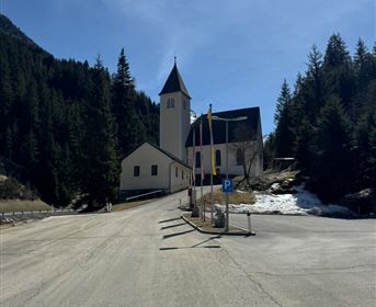 Eine ruhige Straße führt zu einer Kirche in den Bergen. Im Hintergrund sind hohe Bäume zu sehen und der Himmel ist klar.