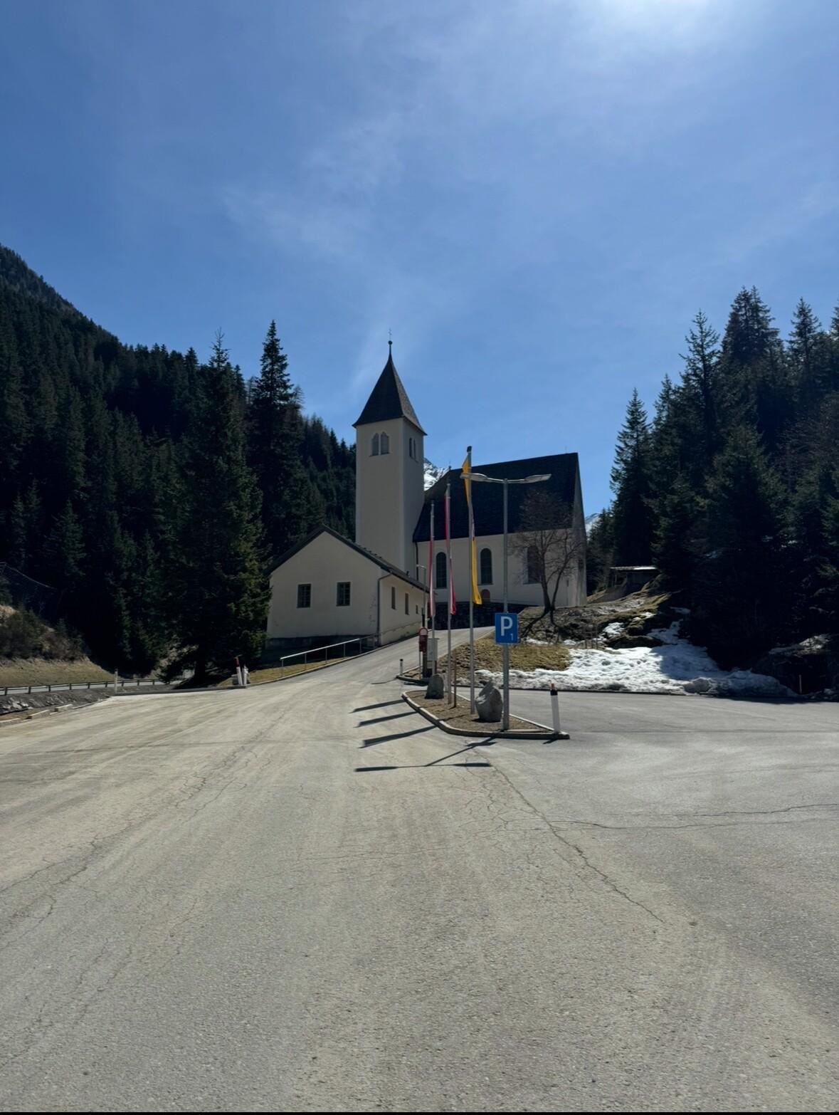 Eine ruhige Straße führt zu einer Kirche in den Bergen. Im Hintergrund sind hohe Bäume zu sehen und der Himmel ist klar.