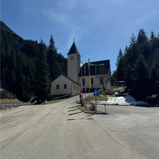 Eine ruhige Straße führt zu einer Kirche in den Bergen. Im Hintergrund sind hohe Bäume zu sehen und der Himmel ist klar.