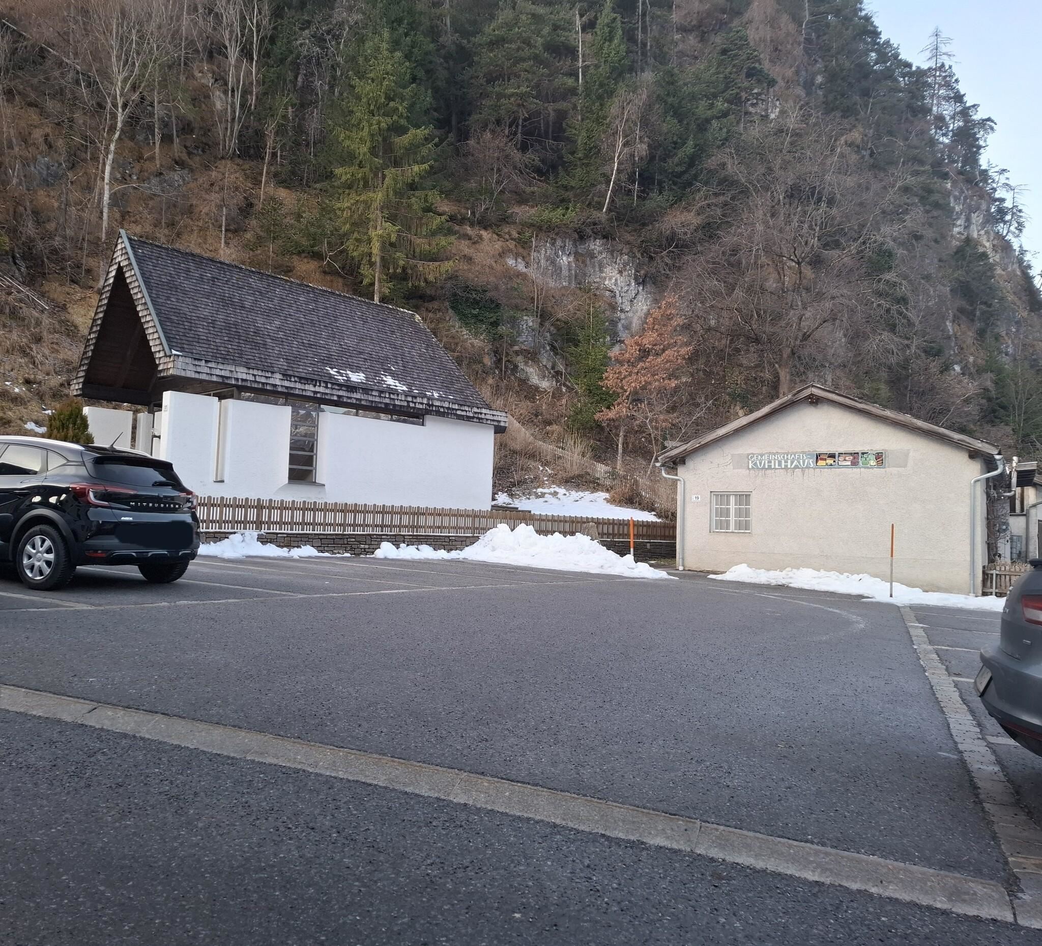 Eine ruhige Winterlandschaft mit schneebedecktem Boden. Im Hintergrund sind zwei Gebäude zu sehen, umgeben von Bäumen.