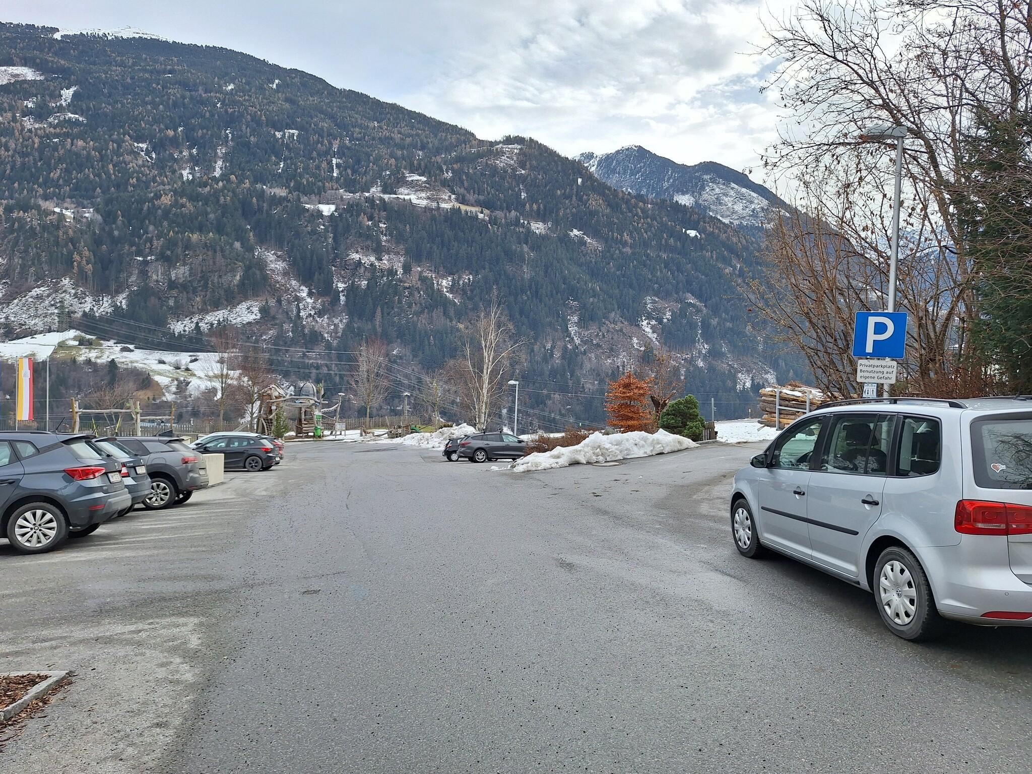 A quiet parking area with cars and snow-covered mountains in the background. The sky is cloudy and there is a cold atmosphere.