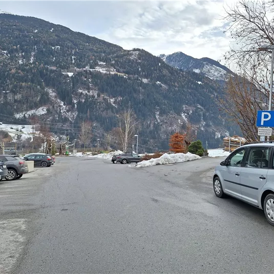 A quiet parking area with cars and snow-covered mountains in the background. The sky is cloudy and there is a cold atmosphere.