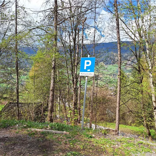 Ein Parkplatz in einer grünen, bewaldeten Umgebung. Im Hintergrund sind sanfte Hügel und ein blauer Himmel zu sehen.
