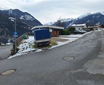 A snow-covered road in the mountains with some houses in the background. A motorhome is parked by the roadside near a parking lot.