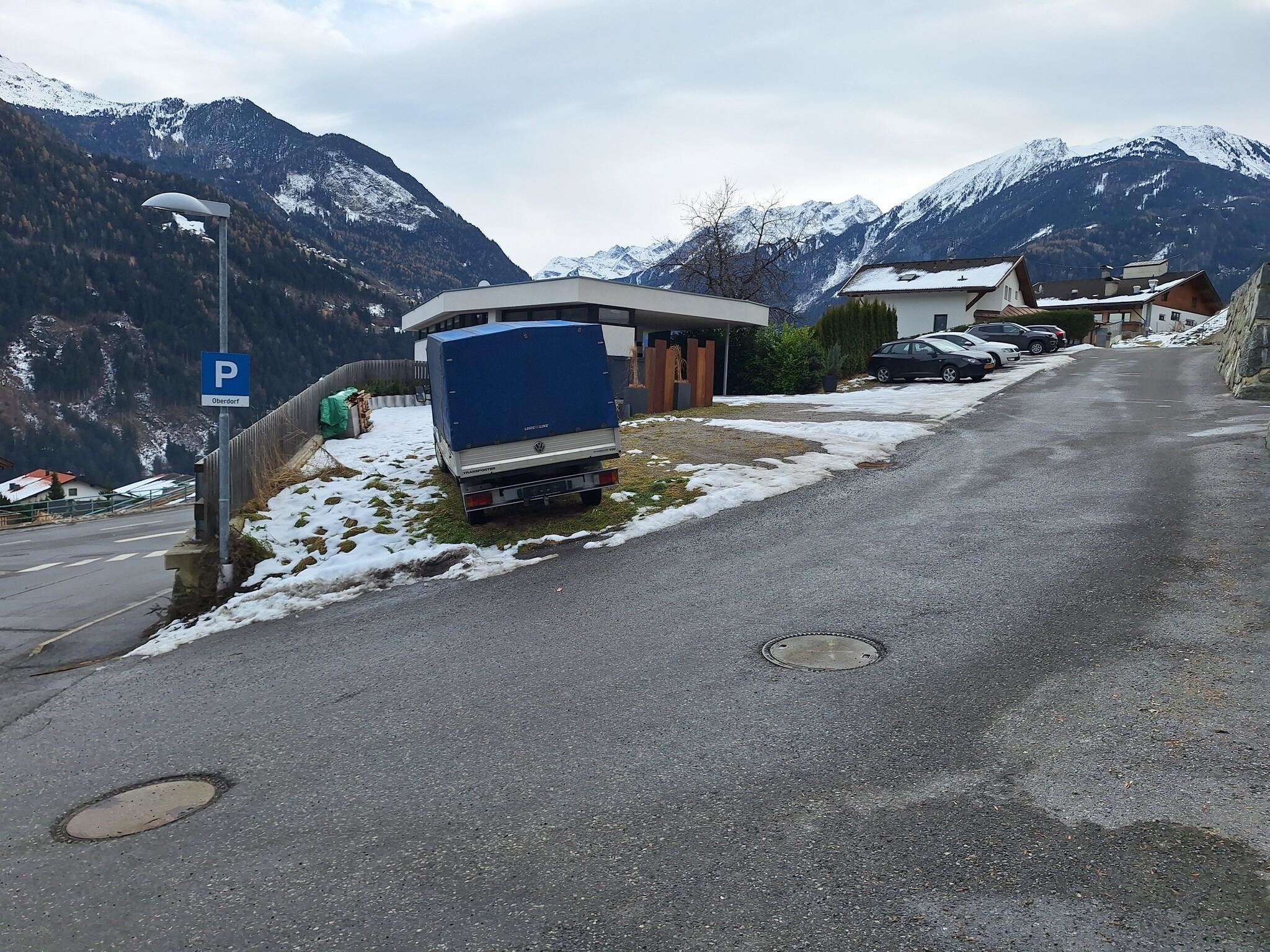 Eine schneebedeckte Straße in den Bergen mit einigen Häusern im Hintergrund. Ein Wohnmobil steht am Straßenrand in der Nähe eines Parkplatzes.