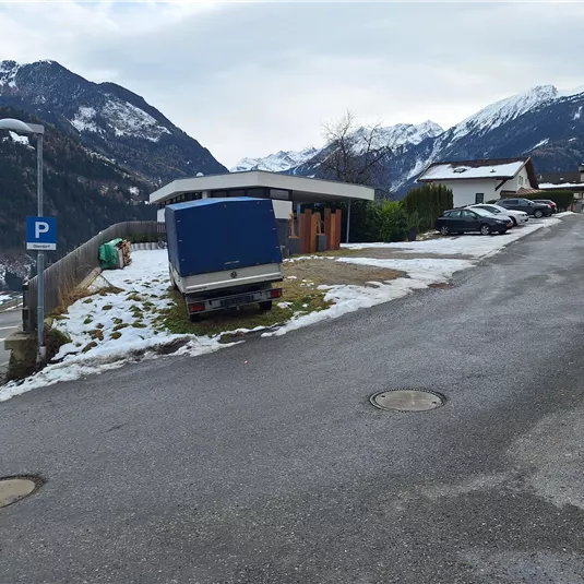 Eine schneebedeckte Straße in den Bergen mit einigen Häusern im Hintergrund. Ein Wohnmobil steht am Straßenrand in der Nähe eines Parkplatzes.