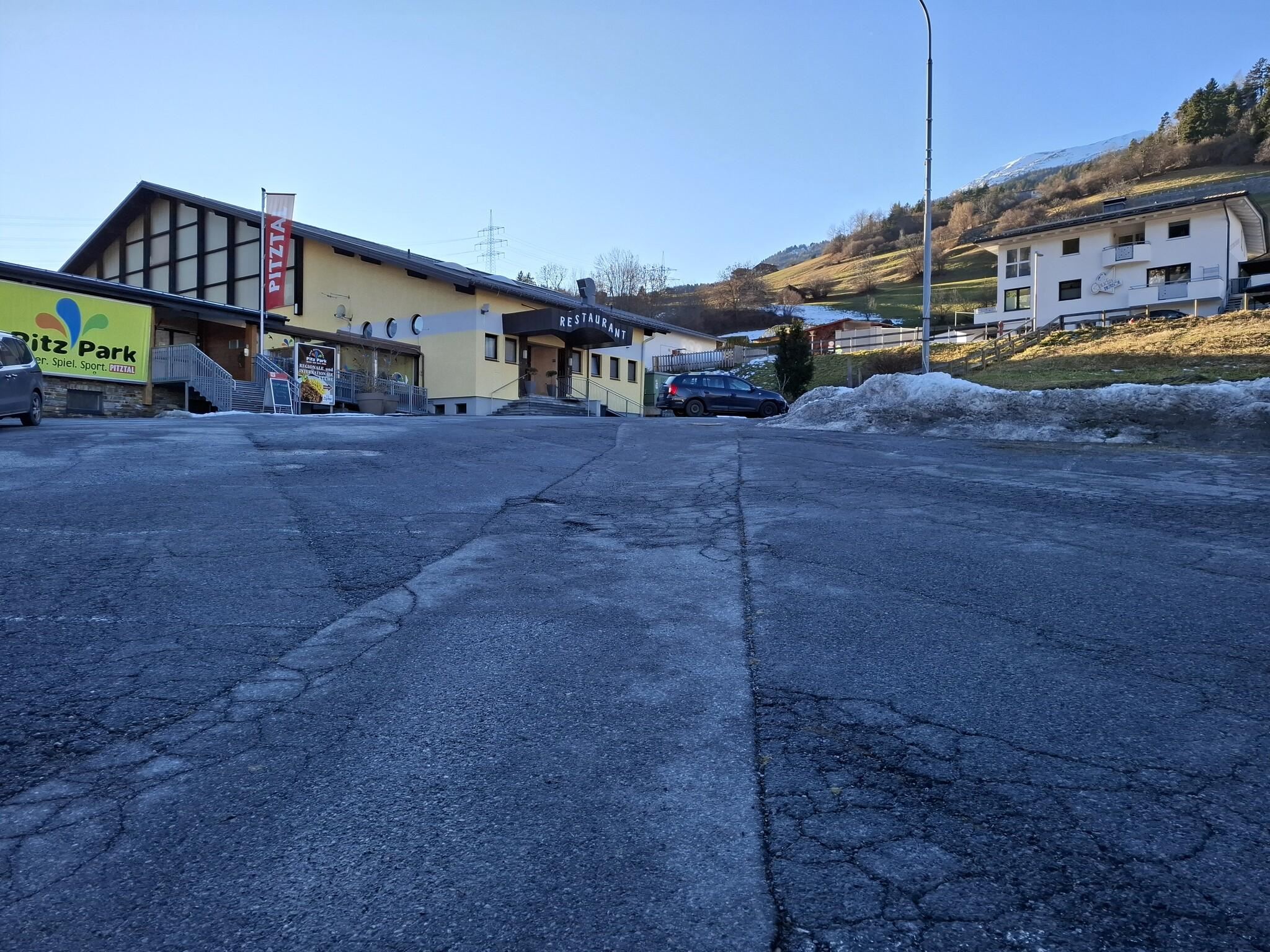 Eine ruhige Straße mit einem Hotel und einigen Wohngebäuden im Hintergrund. Im Vordergrund ist der Asphalt sichtbar, mit noch etwas Schnee an den Rändern.