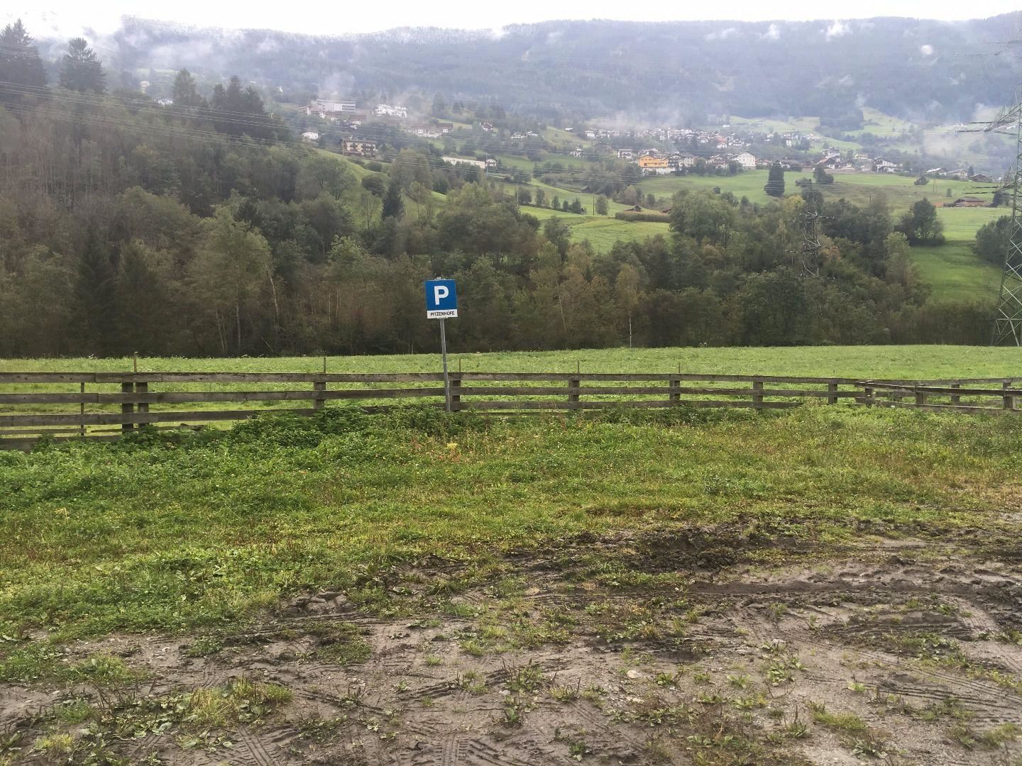 Eine grüne Wiese mit einem Parkplatzschild. Im Hintergrund sieht man sanfte Hügel und einen kleinen Ort.