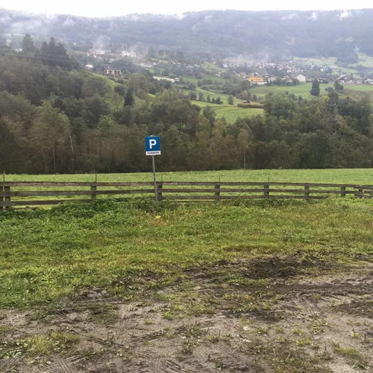 Eine grüne Wiese mit einem Parkplatzschild. Im Hintergrund sieht man sanfte Hügel und einen kleinen Ort.