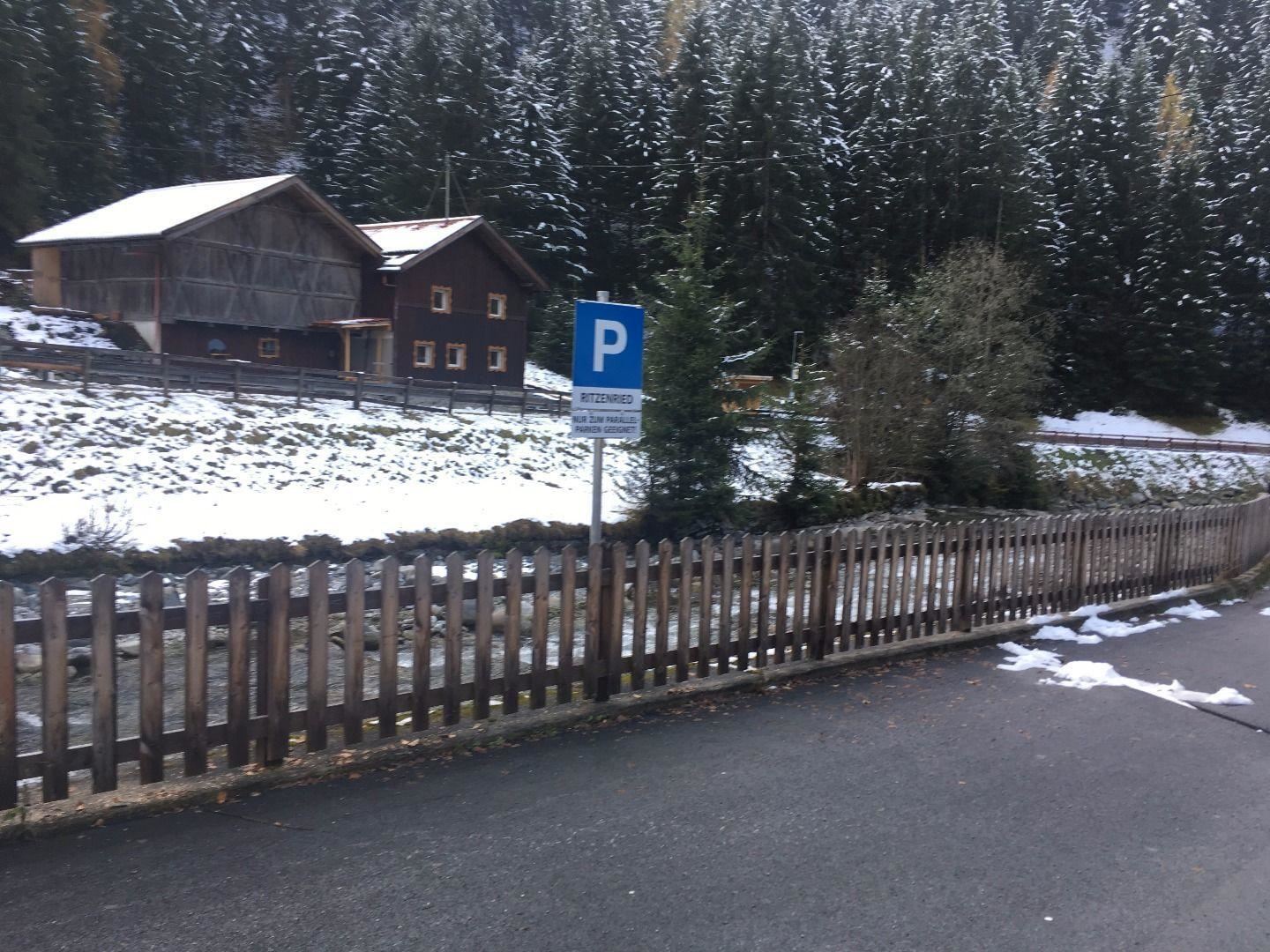 A winter landscape with a parking sign. In the background, traditional wooden houses and snowy trees can be seen.