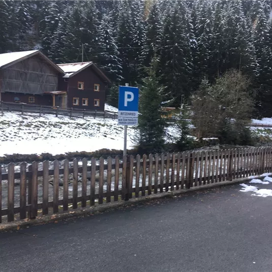 A winter landscape with a parking sign. In the background, traditional wooden houses and snowy trees can be seen.
