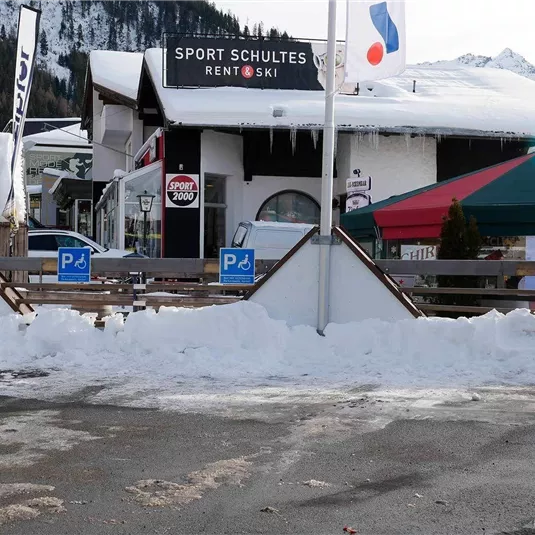 Ein Parkplatz vor einem Gebäude mit dem Namen "Sport Schultes" und schneebedeckten Flächen. Im Hintergrund sind Berge und ein klarer Himmel zu sehen.