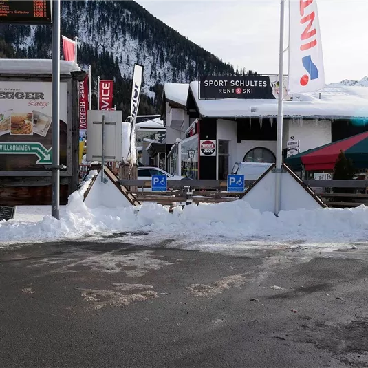 Ein schneebedeckter Platz mit Restaurants und Geschäften in den Bergen. Im Hintergrund sind die schneebedeckten Gipfel sichtbar.