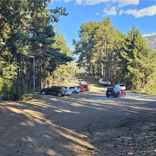 Ein Parkplatz in einem Waldgebiet mit einigen Autos. Im Hintergrund sind Bäume und Berge zu sehen.