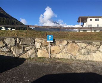 Ein Parkplatz mit einem blauen P-Schild steht an einer Steinmauer. Im Hintergrund sind Berge und ein klarer Himmel zu sehen.