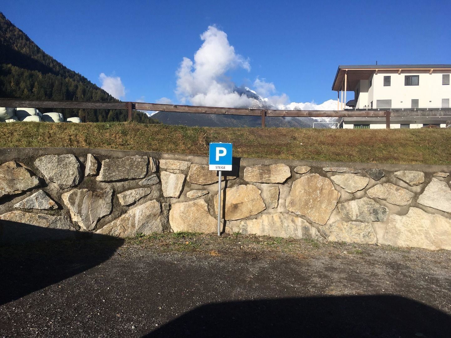 A parking lot with a blue P sign is located by a stone wall. In the background, mountains and a clear sky are visible.