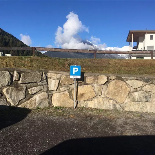 A parking lot with a blue P sign is located by a stone wall. In the background, mountains and a clear sky are visible.
