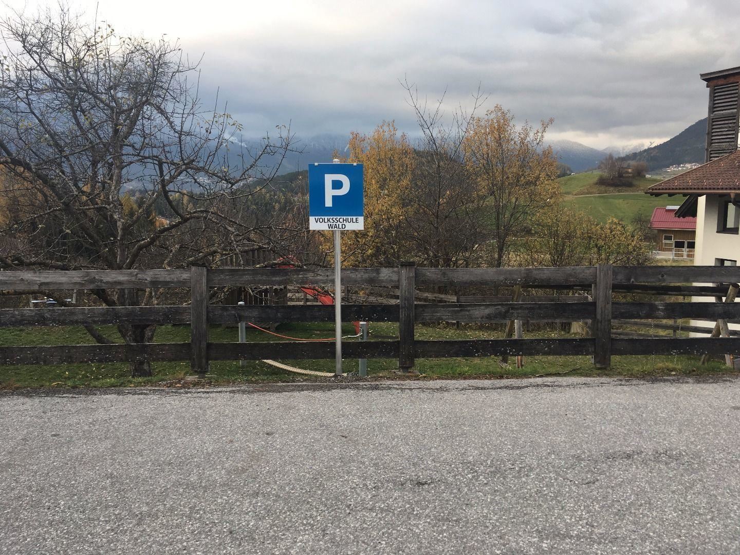 A parking sign stands by a road, surrounded by a wooden fence. In the background, there are trees and hilly landscapes.